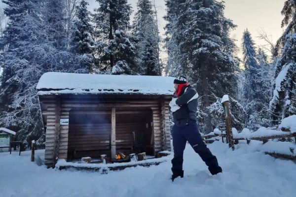 Homem de perfil olhando para um abrigo de madeira no meio de uma floresta nevada.