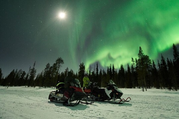 Duas pessoas curtindo a noite de neve com a aurora boreal e motos de neve