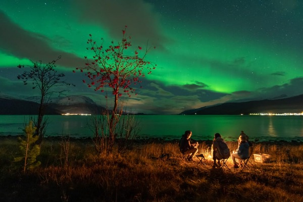 Três pessoas fazendo uma fogueira sob a Aurora Boreal