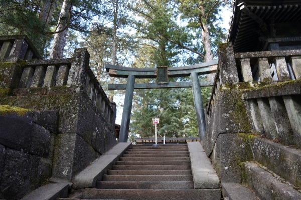 Escadaria de pedra e torii de bronze na excursão a Nikko saindo de Tóquio Escadas de pedra cercadas por muros antigos que levam a um grande torii de bronze em um ambiente arborizado.