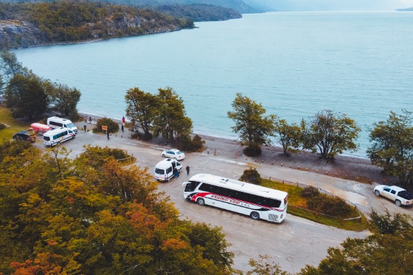 Ônibus turísticos no Parque Nacional da Terra do Fogo. Vista aérea de um estacionamento próximo a um grande lago no Parque Nacional da Terra do Fogo, com vários ônibus turísticos brancos e vans estacionados no local, cercados pela folhagem de outono.