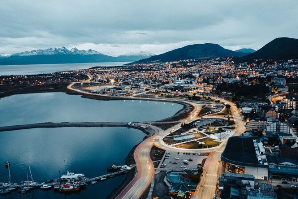 Nachtansicht der Stadt Ushuaia in Argentinien aus der Vogelperspektive. Eine Luftaufnahme von Ushuaia in Argentinien in der Abenddämmerung, mit den Lichtern der Stadt, der mit Booten übersäten Bucht und den Bergen im Hintergrund unter einem bewölkten Himmel.