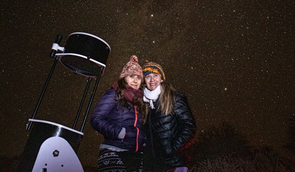 zwei Frauen posieren neben einem Teleskop und mit dem Sternenhimmel im Hintergrund