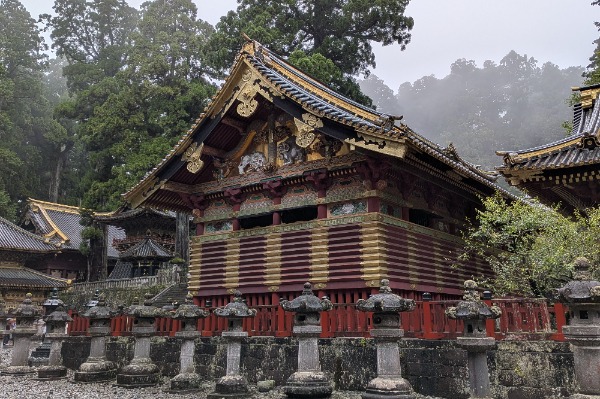 Goldene Details der Heiligen Lagerhäuser auf dem Ausflug von Tokio nach Nikko Traditionelles Gebäude aus rotem und goldenem Holz mit kunstvollen Schnitzereien vor einer Reihe von Steinlaternen.