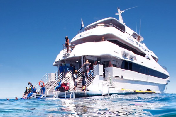 A large, three-level white catamaran with passengers and divers getting ready on the aft deck against the backdrop of the blue sea.