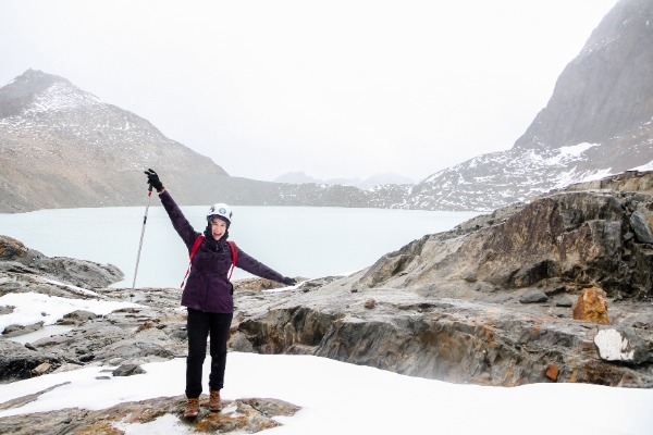 person equipped with crampons and trekking poles posing with open arms with the emerald lagoon in the background
