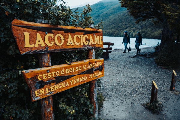 A large carved wooden sign that reads "LAGO ACIGAMI" in yellow letters, with additional text in the Yámana language, located on a trail near the lake's shore, with two hikers walking in the background.