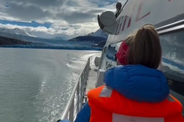 Mujer con gorro y chaleco salvavidas apoyada en el pasamanos exterior contemplando la estela del barco y el glaciar al fondo.