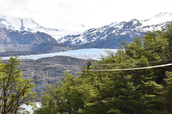 Senderista cruzando el puente colgante con el Glaciar Grey y montañas nevadas al fondo en Torres del Paine