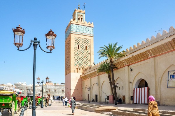 Mezquita y calle tradicional en el centro histórico de Marrakech Calle del centro histórico de Marrakech con mezquita, torre tradicional, palmeras y carruajes en un entorno urbano típico marroquí