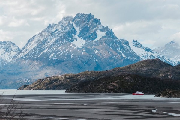 Vue panoramique du bateau naviguant face aux massifs lors de l'excursion « Navigation sur le lac Grey » Vue prise de loin d'un petit bateau rouge naviguant sur le lac Grey, avec en arrière-plan d'imposantes montagnes enneigées.