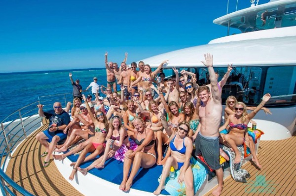Un gruppo di passeggeri che si gode il sole sul ponte del catamarano Un folto gruppo di turisti in costume da bagno che salutano allegramente dal ponte esterno della nave in mare aperto.