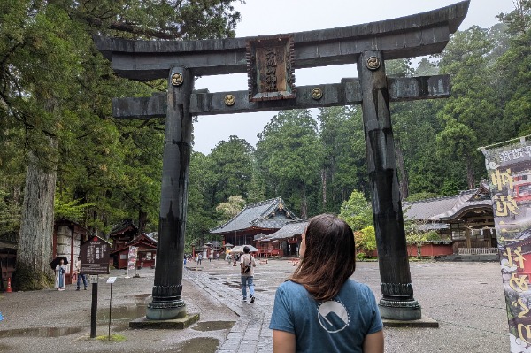 Grande torii in bronzo all’ingresso del santuario durante l’escursione a Nikko da Tokyo Una donna che osserva l'imponente torii in bronzo con gli edifici tradizionali del santuario sullo sfondo in una giornata nuvolosa.